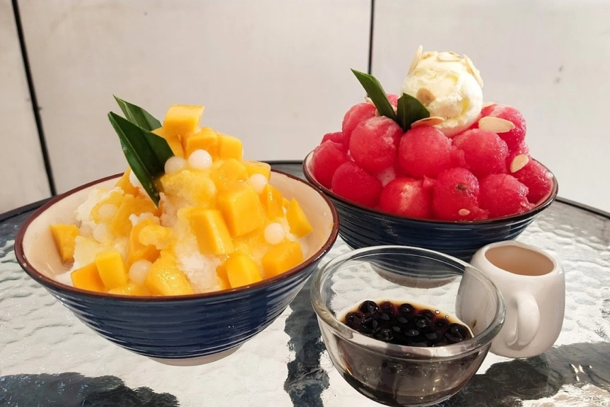 A bowl of bingsu with mango on top and a bowl of bingsu with watermelon balls and a scoop of icecream on a glass table