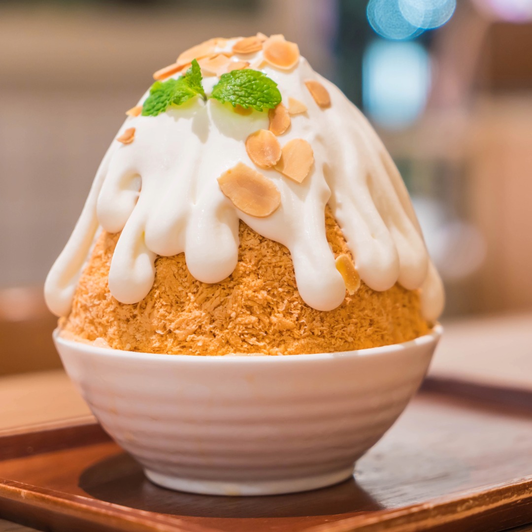 A picture of a bowl of orange colored bingsu topped with a white dripped topping, almond slices, and a garnish of mint